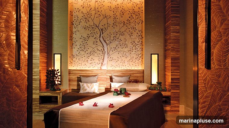 A serene spa treatment room with two massage beds. The walls are decorated with textured panels and a large mural of a tree. Soft lighting from wall sconces and accent lighting creates a calming ambiance. Each bed is prepared with linens, pillows, and decorative elements like flowers and small vases.