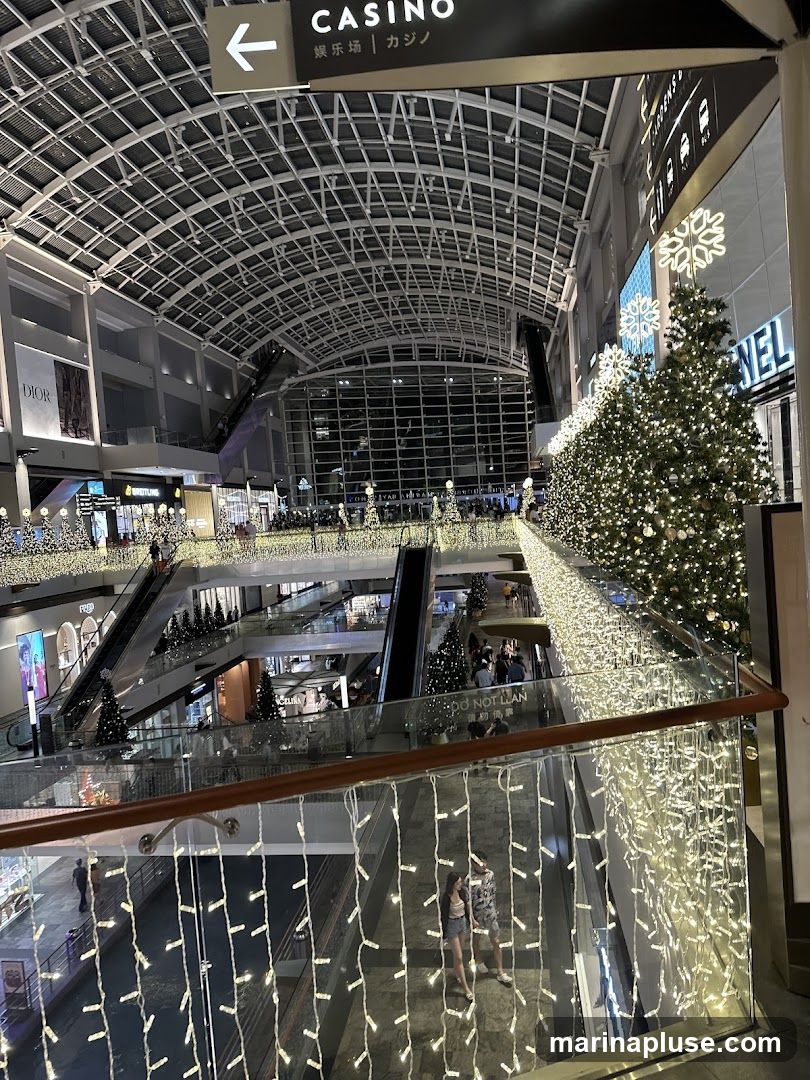 The image shows the interior of a multi-level shopping mall decorated with Christmas lights and trees. Escalators and walkways are visible, with shoppers moving around. Signs for various shops, including Dior and Breitling, can be seen. A sign indicating "CASINO" with an arrow is also present. The overall atmosphere is festive and commercial.