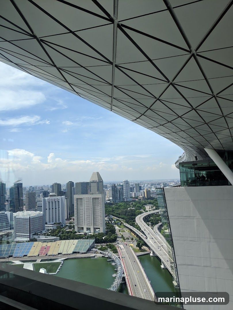 A panoramic view of a bustling city skyline, featuring numerous skyscrapers and a complex network of highways and bridges. The foreground is dominated by the distinctive geometric pattern of a modern architectural structure, possibly a rooftop or canopy, with the sky visible through its triangular panels. Below, a body of water reflects the urban landscape, and a stadium with colorful seating is visible. The scene captures a bright, clear day with scattered clouds.