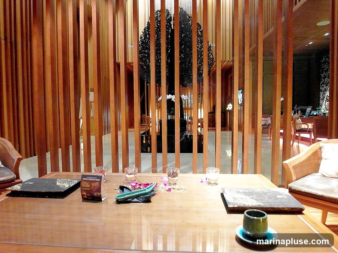 A table is set in a restaurant with wooden slats in the background. On the table, there are two menus, small glasses with what appears to be decorative items, a green ceramic dish, and a cup of tea. The background shows a well-lit interior with more wooden elements, decorative plants, and seating areas.