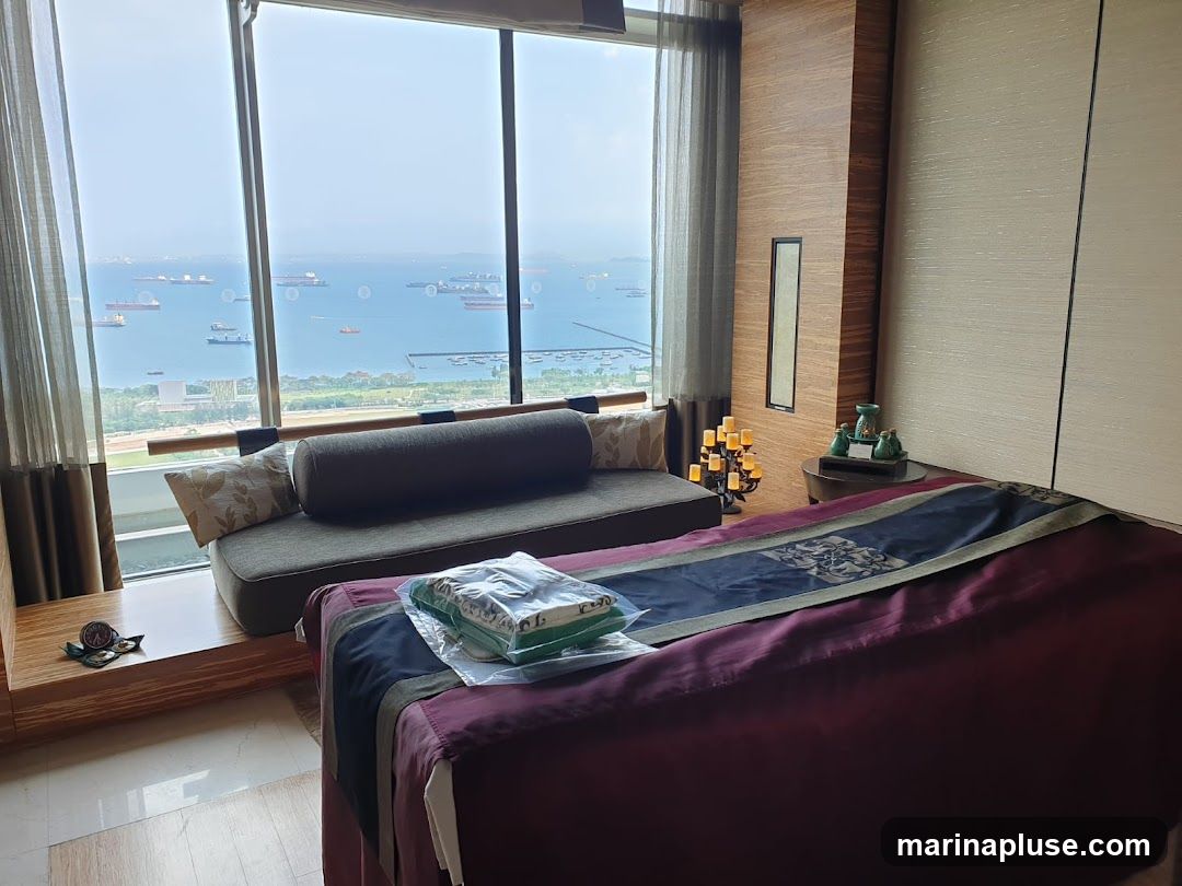 A spa room with a large window overlooking the ocean and numerous ships. The room features a massage bed with a decorative runner, a couch, and decorative items like candles and vases.