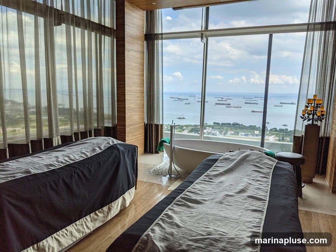 A spa room with two massage tables, a bathtub, and large windows offering a panoramic view of the ocean with many ships. The room has wooden walls and sheer curtains.