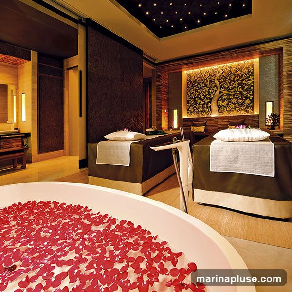 A serene spa suite featuring a large bathtub filled with red rose petals in the foreground. Two massage beds are set up in the background, each with a pillow and white towel. The room is dimly lit with ambient lighting, including decorative lights on the ceiling resembling stars and illuminated panels on the walls. A large, ornate wall art depicting a tree with intricate patterns is a focal point behind the massage beds. To the left, a vanity area with a sink and wooden cabinetry is visible.