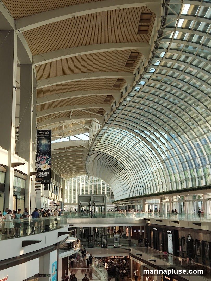 The image shows the interior of a large, modern shopping mall with a distinctive curved glass roof. Multiple levels of shops and walkways are visible, with shoppers browsing and moving around. Natural light streams in through the glass ceiling, illuminating the spacious atrium. A large banner hangs from the ceiling with text that appears to be an advertisement or event promotion.