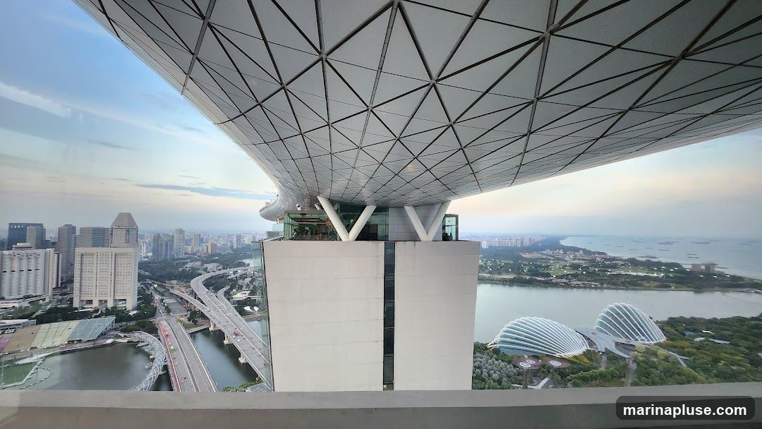 A panoramic view from a high vantage point, likely a skyscraper observation deck, overlooking a city skyline and a body of water. The foreground features the distinctive geometric pattern of the building's roof structure. In the distance, a dense urban landscape with numerous buildings is visible, along with a highway system and a bridge. To the right, the iconic domes of Gardens by the Bay can be seen, along with a coastline and the sea dotted with ships. The sky is overcast with soft, diffused light, suggesting either early morning or late afternoon.