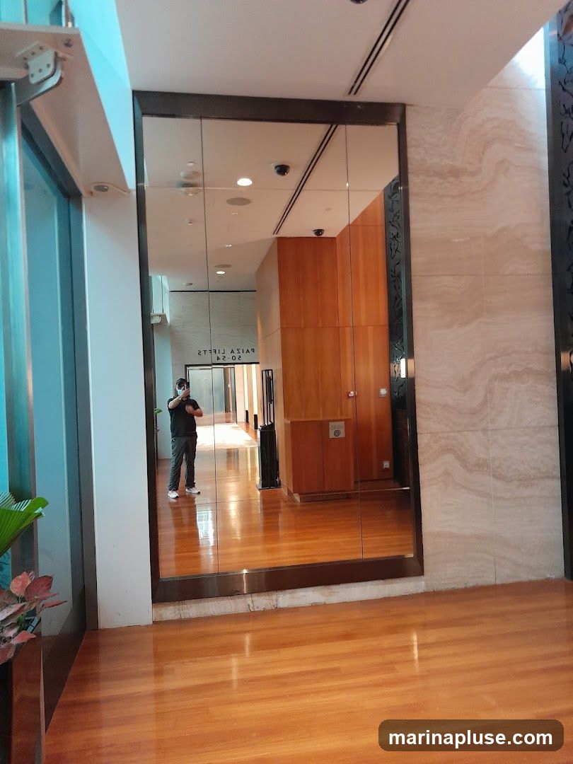A person takes a selfie in a hallway with polished wooden floors and a large mirrored wall. The mirror reflects the hallway, including ceiling lights, security cameras, and wooden paneling on the walls. Text is visible in the reflection, reading "PAIZA LIFTS 02-04".