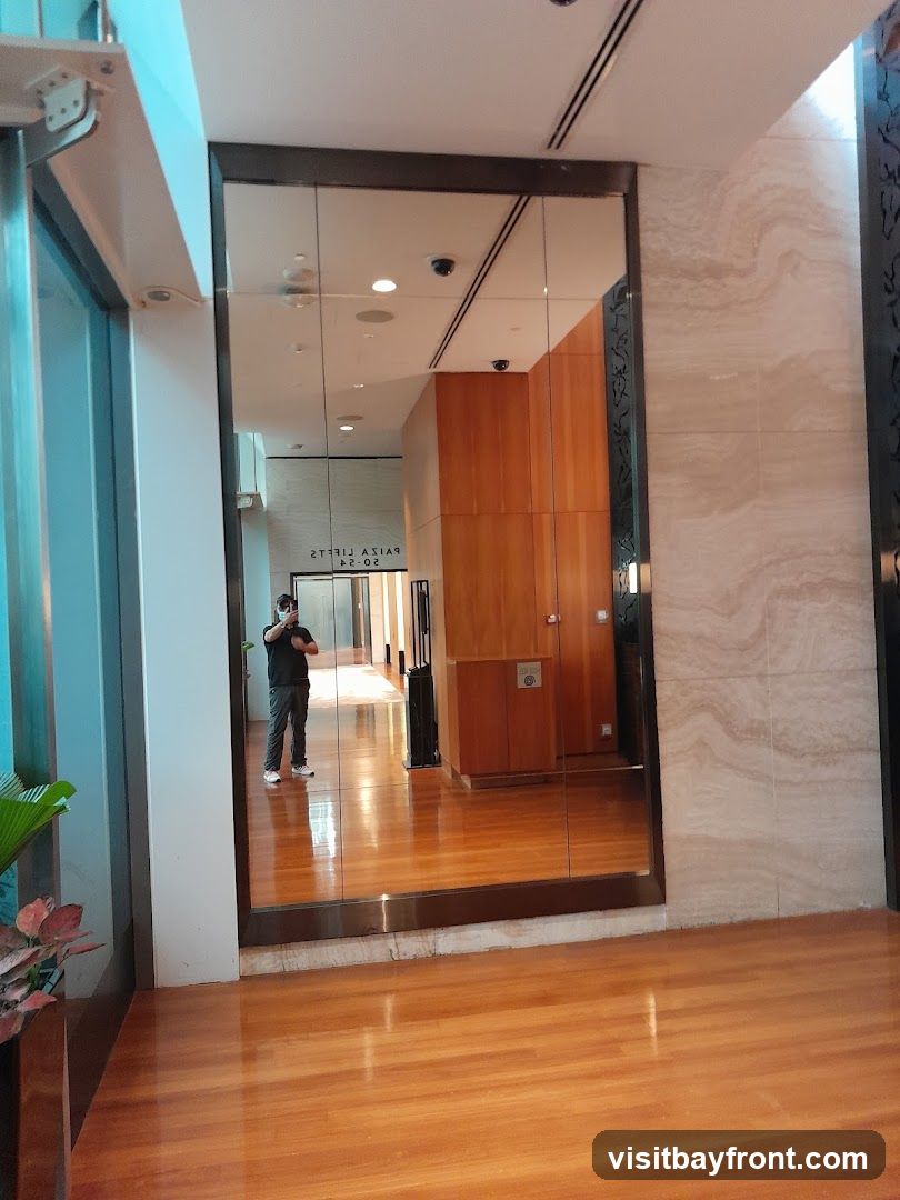 A person takes a selfie in a hallway with polished wooden floors and a large mirrored wall. The mirror reflects the hallway, including ceiling lights, security cameras, and wooden paneling on the walls. Text is visible in the reflection, reading "PAIZA LIFTS 02-04".