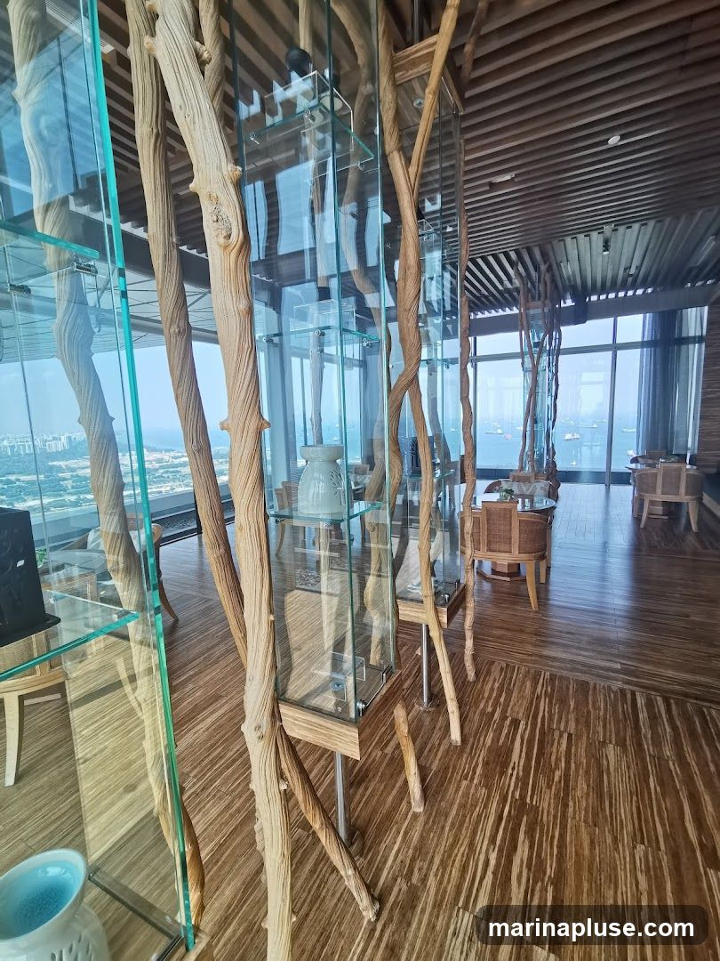 The image shows the interior of a room with a modern, naturalistic design. Tall, slender glass display cases with wooden frames are arranged throughout the space, showcasing decorative items. These cases are intertwined with natural, twisted branches, adding an organic element to the decor. The flooring is made of polished bamboo with a distinctive striped pattern. In the background, large windows offer a panoramic view of the ocean and distant city skyline. Seating areas with wicker chairs and tables are visible, suggesting a lounge or dining area. The overall ambiance is sophisticated and serene, with a focus on natural materials and scenic views.