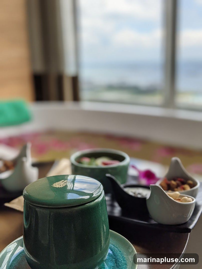 A close-up view of a spa setting with various small bowls of snacks and a lidded container, likely part of a spa treatment experience. In the background, a bathtub filled with water and flower petals is visible, with a window offering a blurred view of the outdoors and a body of water.