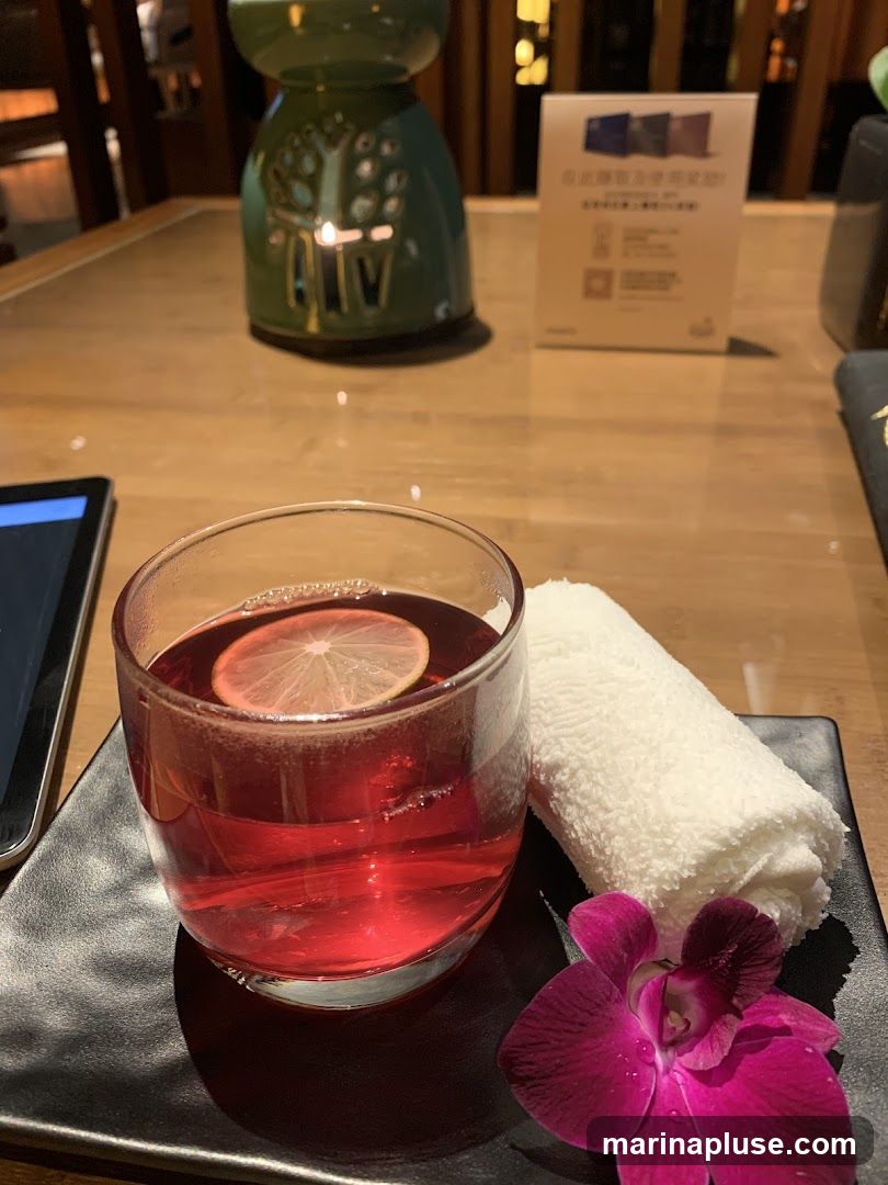 A glass of red beverage with a slice of lemon and a rolled white towel are placed on a black square plate. A purple orchid is positioned in the lower right corner of the plate. In the background, a green decorative object with a lit candle inside sits on a wooden table. A small card with text and QR codes is also visible on the table.