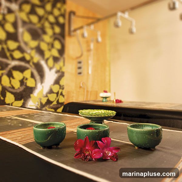 A spa treatment room with a massage table, shower area, and decorative elements. In the foreground, there are three small green bowls filled with water and red petals, with two red orchids placed in front of them. A green decorative dish with sliced cucumbers is visible on the table behind the bowls. The background shows a showerhead system and a wall with a decorative pattern of yellow leaves on a dark background.