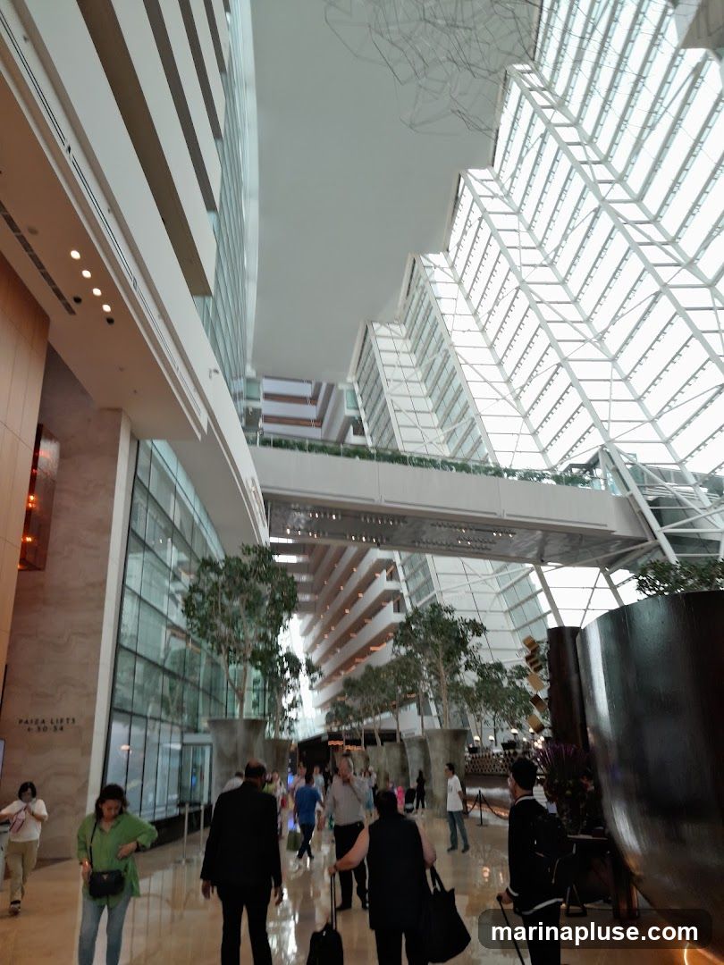 A wide-angle shot of a grand hotel atrium with a distinctive geometric glass ceiling. People are seen walking through the spacious lobby, which features tall indoor trees and modern architectural elements. The interior design is sleek and contemporary, with a focus on natural light and open space.