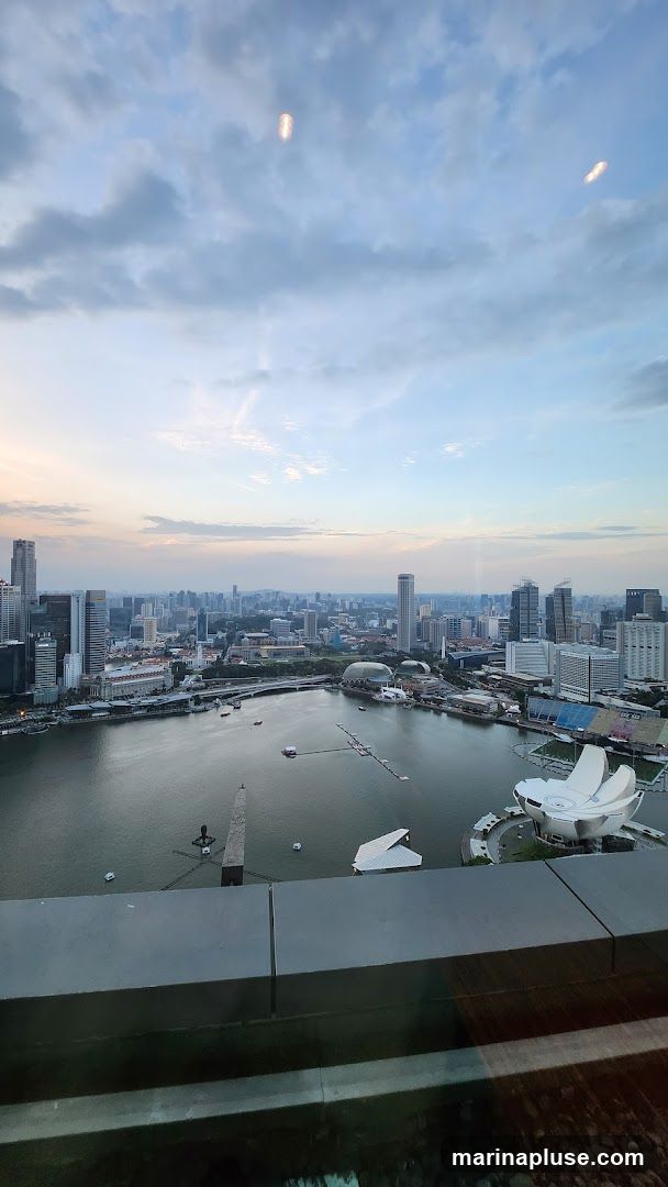 A panoramic view of a city skyline at dusk, with a body of water in the foreground. Tall buildings dominate the landscape, and the sky is filled with clouds, with hints of sunset colors. Lights are beginning to appear in the buildings and on the water.