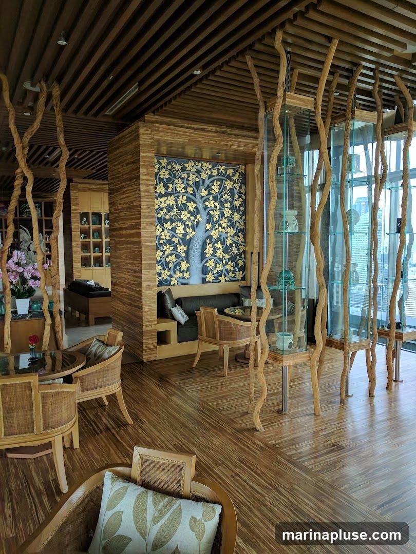The image shows the interior of a lounge area with a natural and organic design. The walls and ceiling are made of wood, with a slatted wooden ceiling. Decorative branches are used as structural elements and accents throughout the space. There are several seating areas with wicker chairs and sofas, some with patterned cushions. A large wall mural of a tree with golden leaves is a focal point. Glass display cabinets with decorative items are also visible. The flooring is made of polished wood.