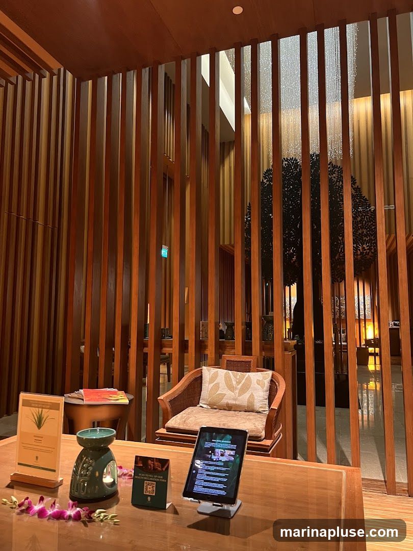 A spa reception area with a wooden slatted screen, a comfortable armchair, and a wooden counter. On the counter, there is a tablet displaying a form, a QR code, a decorative oil burner, and a pamphlet. Orchids are placed on the counter. In the background, a decorative tree and a water feature are visible.