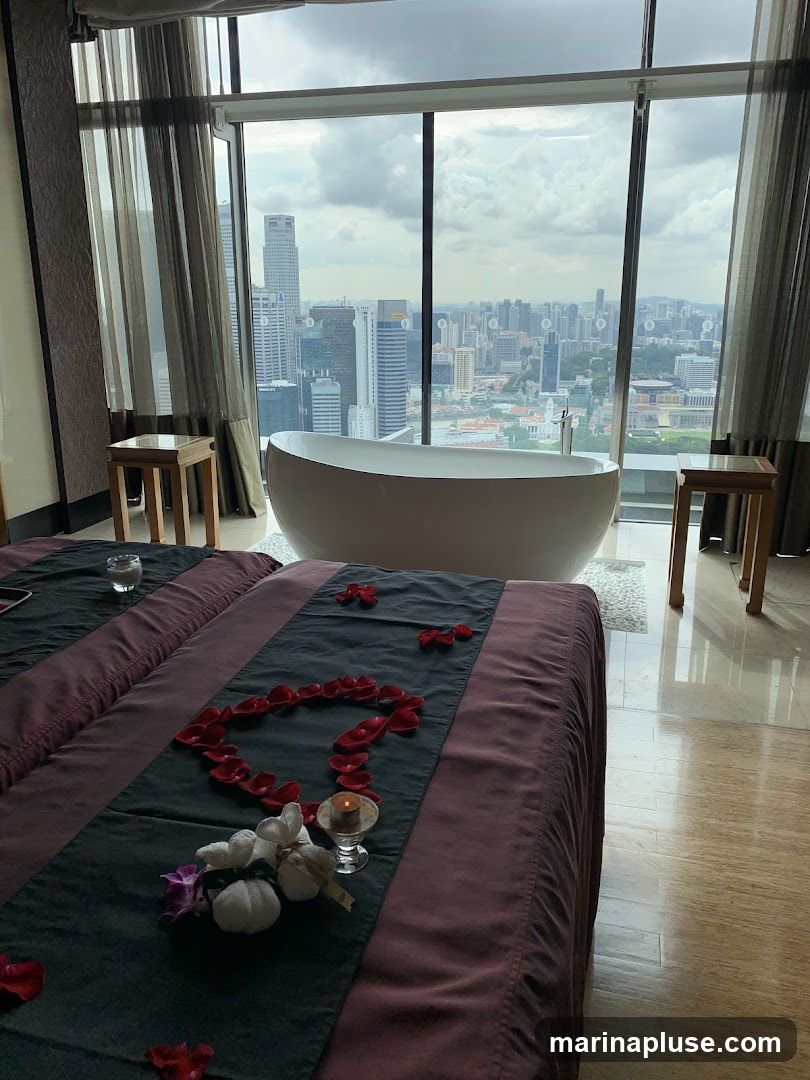 A spa room with two massage beds decorated with rose petals and a heart shape. A bathtub is positioned in front of a large window with a panoramic view of a city skyline. Two small wooden tables are placed on either side of the bathtub.