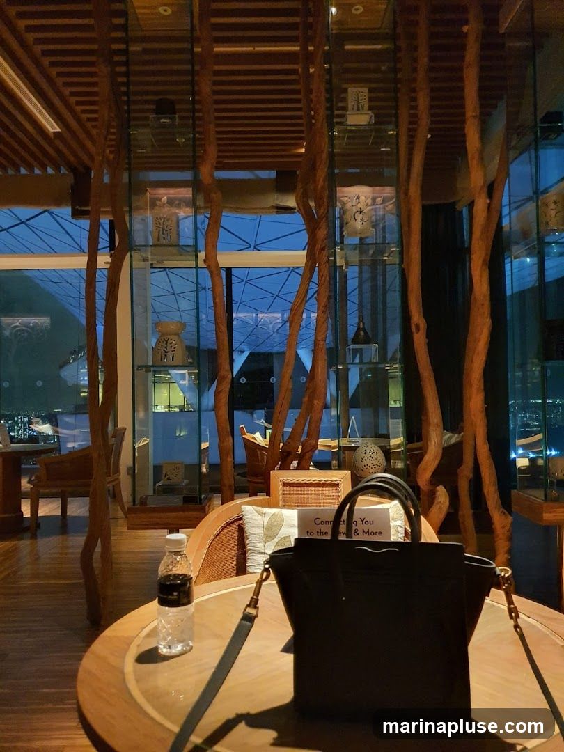 The image shows the interior of a lounge or restaurant with a large round wooden table in the foreground. On the table, there is a bottle of water and a black handbag. Behind the table, there are chairs and display shelves with decorative items. The background features large windows with a view of a city at night. The decor includes natural elements like wooden branches and a wooden slatted ceiling. A small sign on a cushion reads "Connecting You to the New & More".