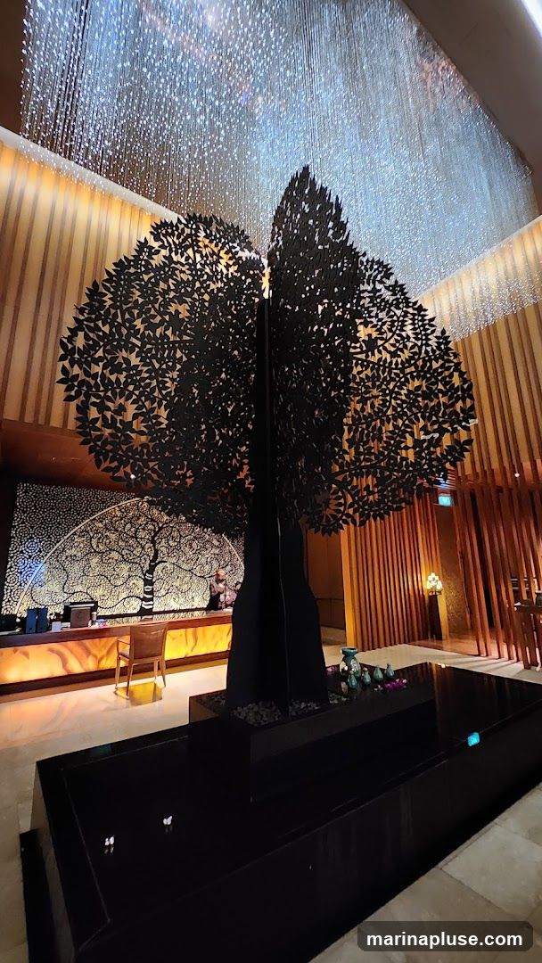 A large, black, metal tree sculpture stands in the center of a modern lobby. The sculpture is intricately detailed with many leaves. Above the sculpture, a dazzling light fixture hangs, resembling a cascade of tiny lights. In the background, a reception desk is visible with a decorative mural of a tree behind it. The lobby features warm wood paneling and ambient lighting.