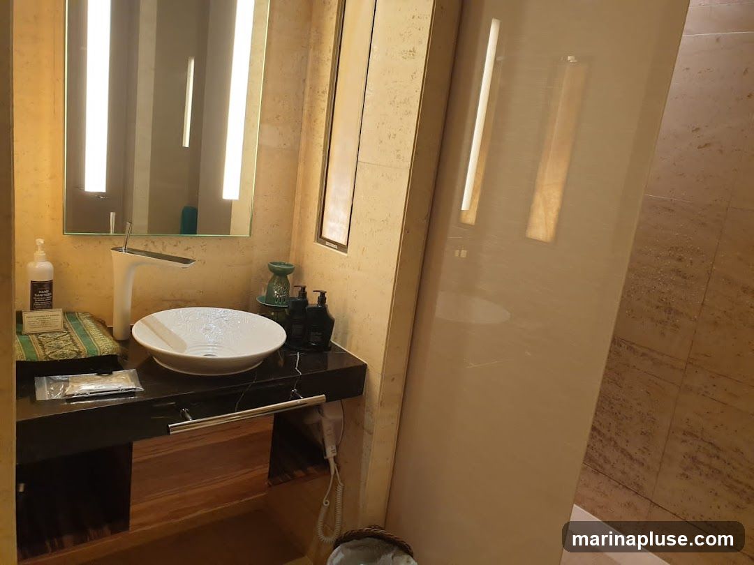 A modern bathroom vanity with a white vessel sink, a sleek chrome faucet, and a black countertop. The vanity features wooden drawers and is illuminated by vertical lights integrated into the mirror. Various toiletries, including hand sanitizer and soap dispensers, are arranged on the counter. The walls are tiled with a neutral stone pattern, and a glass shower enclosure is visible to the right.