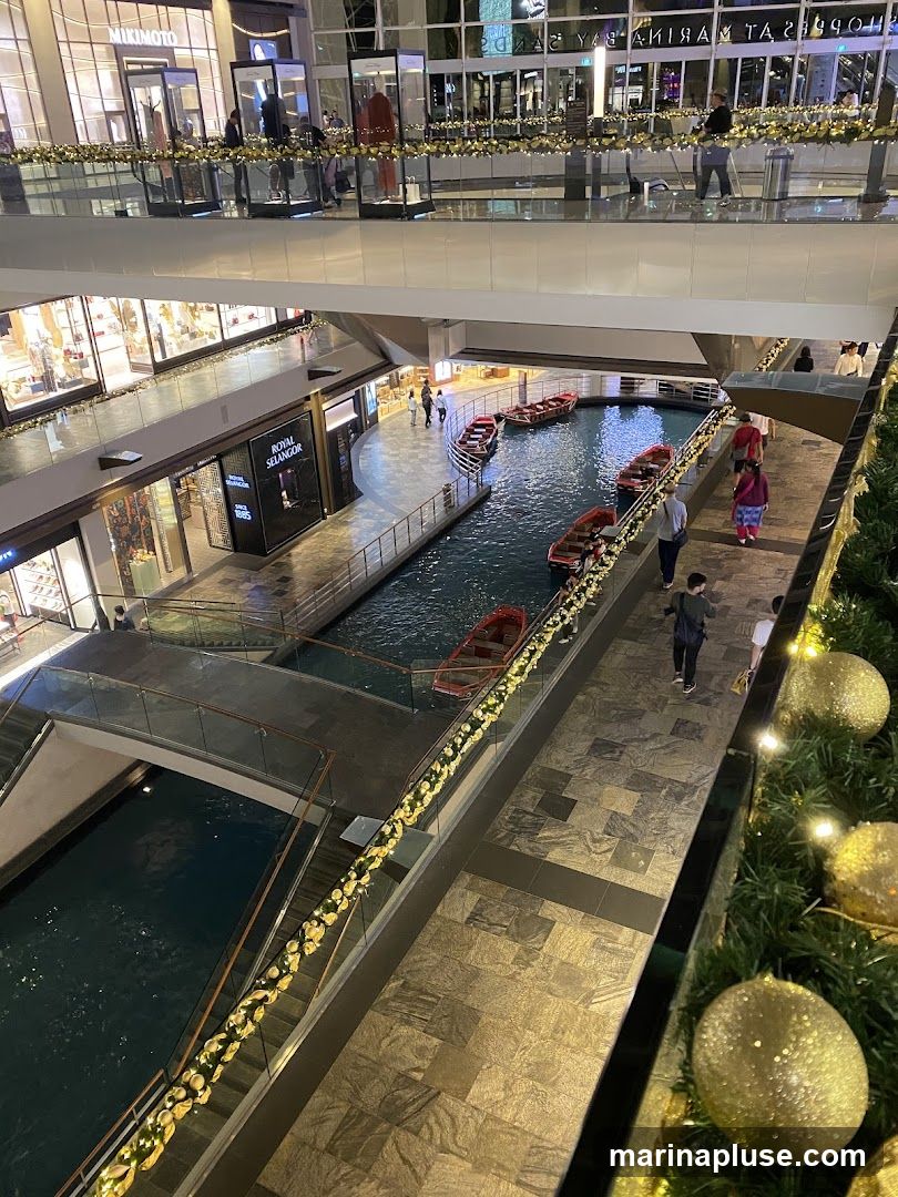 An interior view of a multi-level shopping mall featuring a canal with small boats. Shops are visible on different levels, with displays and signage. People are walking along the walkways and near the canal. Festive decorations, including garlands and large golden ornaments, are present, suggesting a holiday season.