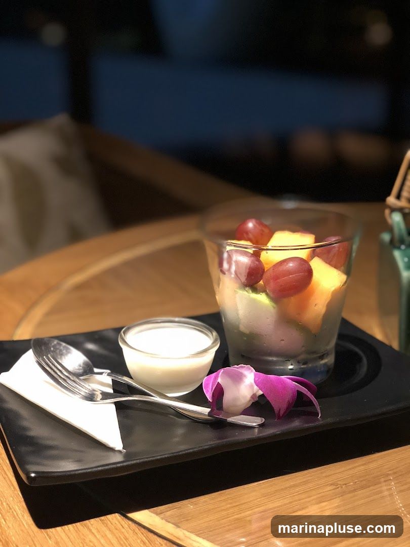 A glass of fruit salad containing grapes, melon, and possibly kiwi, served with a small dish of cream or yogurt. A purple orchid and a spoon and fork are placed on a black rectangular plate. The setting appears to be indoors, possibly a restaurant, with a wooden table and blurred background.