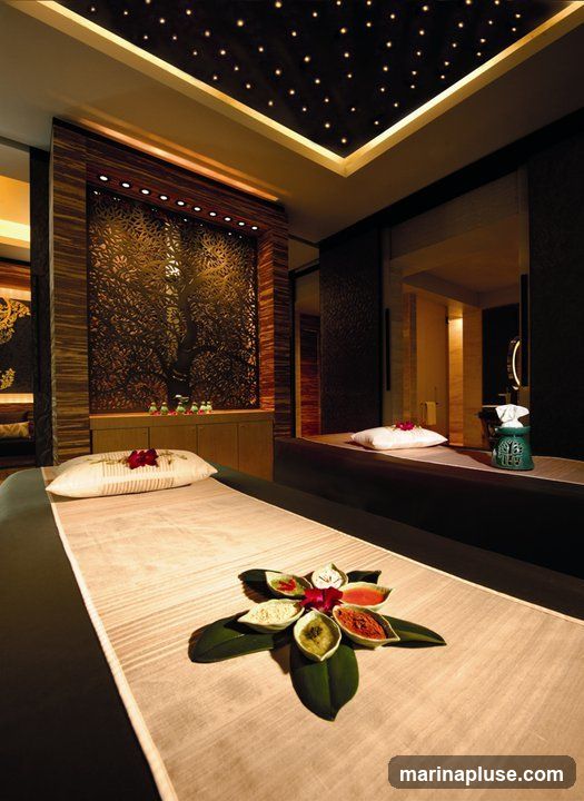 A luxurious spa treatment room with a massage bed prepared for a client. The bed is covered with a light-colored mat and a pillow adorned with a red flower. A decorative arrangement of small bowls containing colorful powders and pastes, possibly for treatments, is placed on the mat. The room features elegant decor, including a ceiling with fiber optic lights resembling stars, wooden paneling, and a decorative screen with a tree motif. A glimpse into an adjacent area shows a sink and a tissue dispenser.