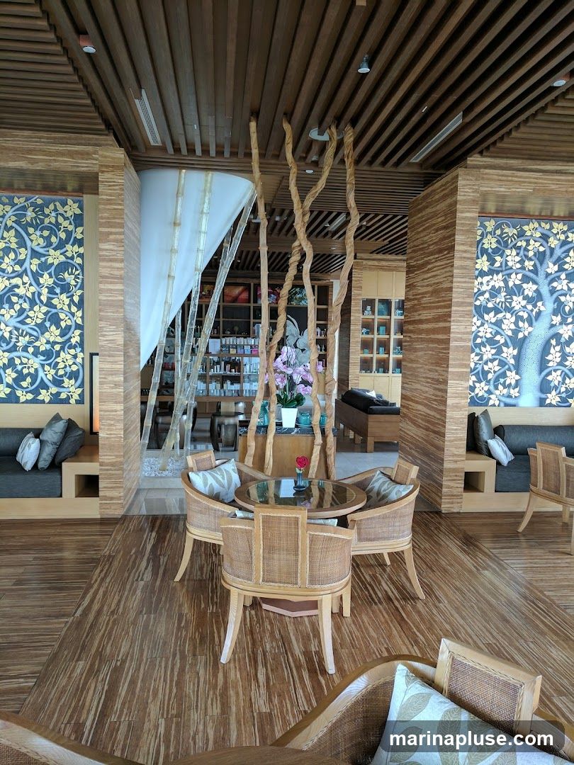 A serene spa waiting area with a central seating arrangement of wicker chairs around a glass-top table. The room features natural wood elements, including a slatted ceiling and bamboo-patterned flooring. Decorative blue and gold wall hangings adorn the walls, and a sheer white curtain with bamboo accents adds a touch of elegance. In the background, shelves display various products, suggesting a retail or treatment area.