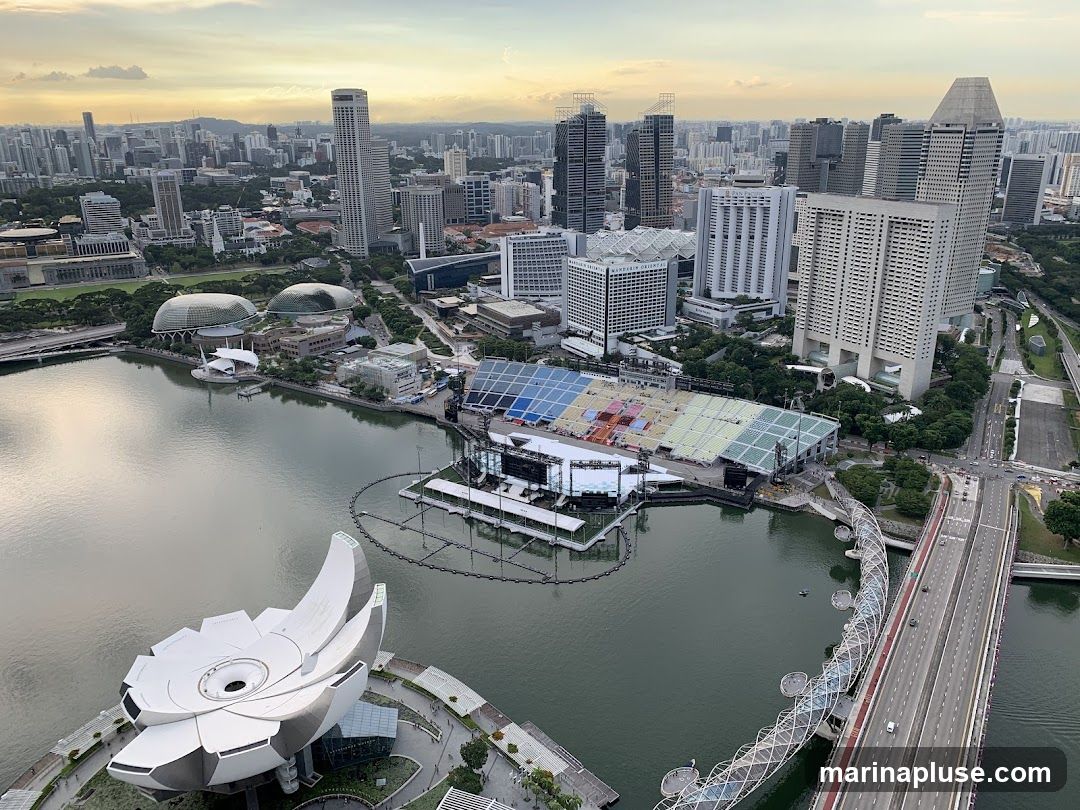 An aerial view of Singapore's Marina Bay area at dusk. The image showcases the city's modern architecture, including skyscrapers, the Marina Bay Sands resort, and the ArtScience Museum. The floating stadium and the Helix Bridge are also visible, with the calm waters of the bay reflecting the sky.