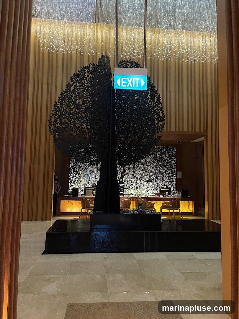The image shows a hotel lobby with a large, decorative black tree sculpture in the center. Behind the tree is a reception desk with warm lighting and chairs. An illuminated green exit sign is visible above the tree. The walls are decorated with vertical wooden panels and a patterned mural. The floor is tiled.