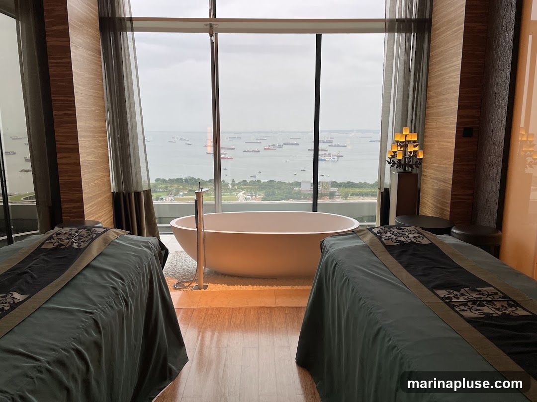 A luxurious spa room with two massage beds facing a large window with a view of the ocean and numerous ships. A freestanding bathtub is positioned in front of the window. The room is decorated with elegant curtains and ambient lighting.