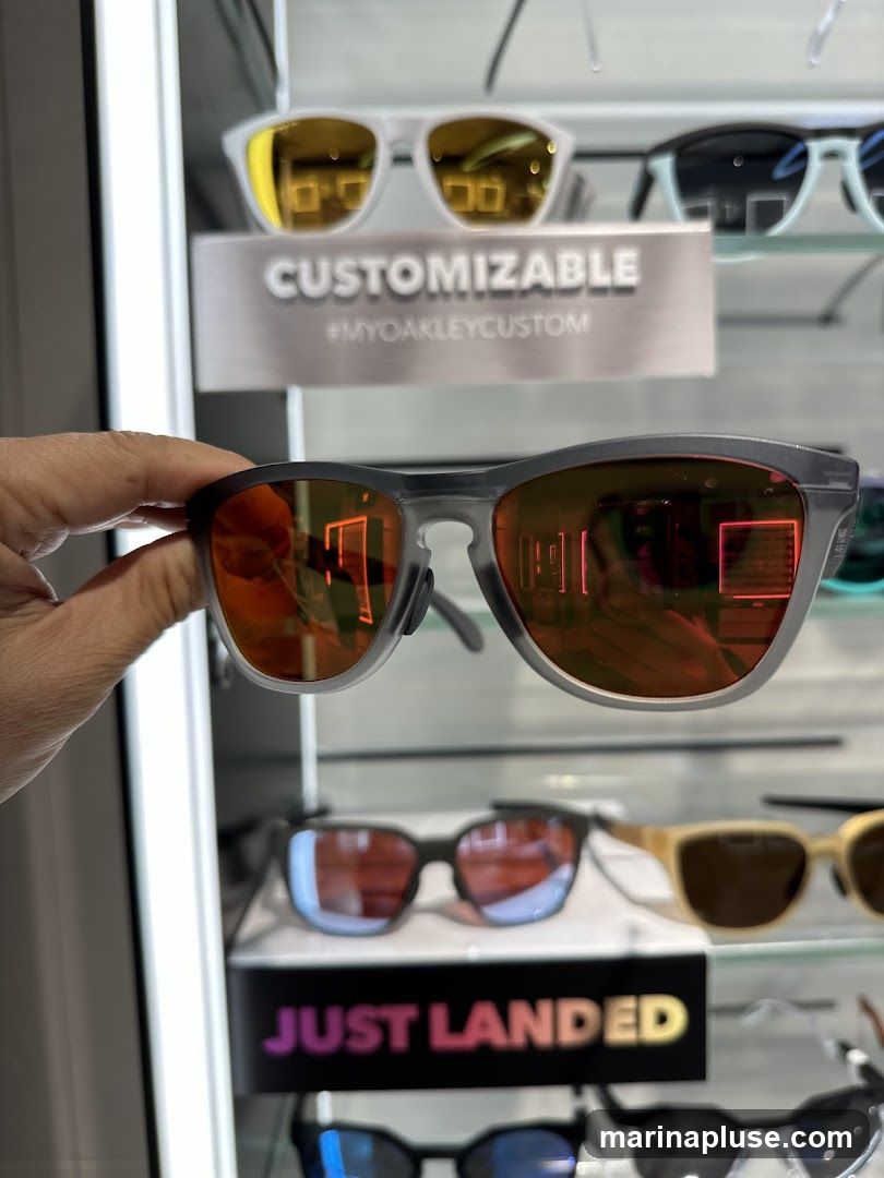 A hand holds a pair of grey Oakley sunglasses with orange lenses. In the background, a display case shows various other sunglasses, including a white pair with gold lenses and a light blue pair. A sign above the white sunglasses reads "CUSTOMIZABLE #MYOAKLEYCUSTOM". Below the grey sunglasses, another sign reads "JUST LANDED" with colorful lettering.