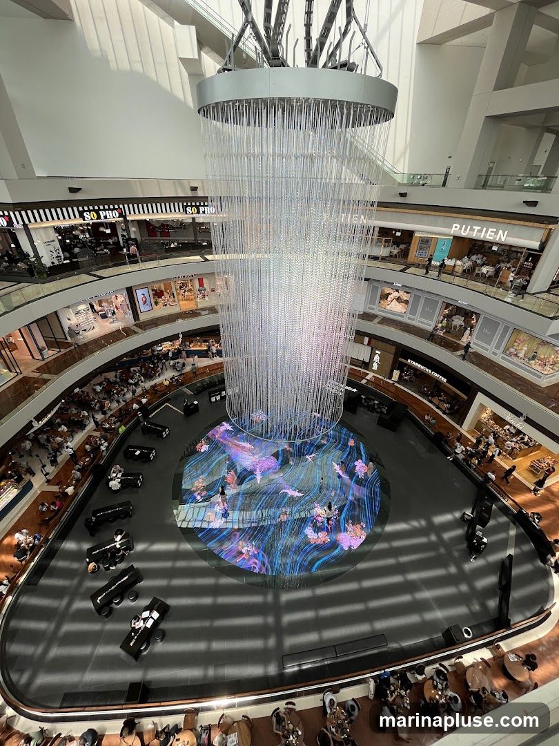 An aerial view of a multi-level indoor atrium featuring a large, ornate chandelier suspended from the ceiling. Below the chandelier, a circular digital display on the floor shows a vibrant, animated scene with aquatic or floral motifs. Several levels of shops and restaurants surround the atrium, with patrons visible in various seating areas and walking around. Some visible shop names include "SO PHO", "PUTIEN", "STARBUCKS COFFEE", and "DIOR".