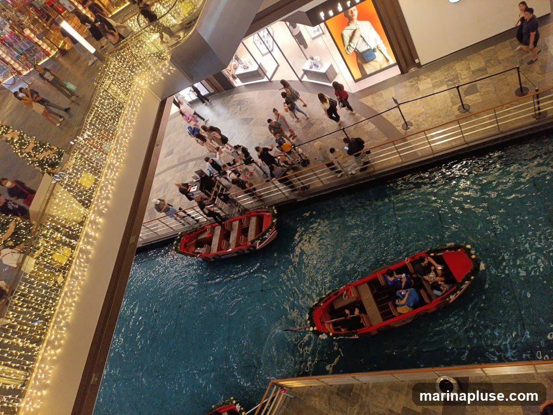 An overhead view of an indoor canal with two red boats carrying passengers. Shoppers are visible on the walkway above the canal, some waiting in line. The walkway is lined with shops, one of which has a large display window featuring a model wearing a light-colored outfit. Decorative lights and Christmas trees are also visible along the upper levels of the building.