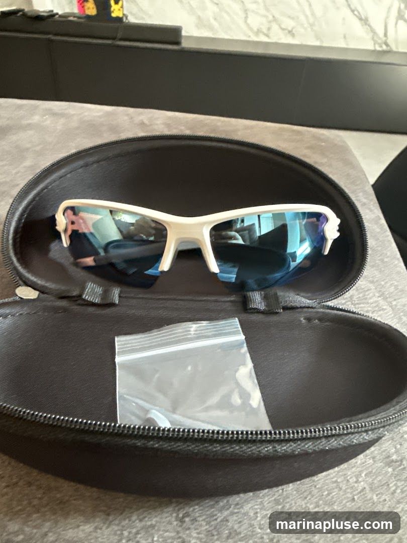 A pair of white sports sunglasses with blue mirrored lenses are open inside a black protective case. A small plastic bag containing what appear to be replacement nose pads or ear tips is also in the case. The case is resting on a textured gray surface, possibly a couch or fabric. In the background, a black shelf or cabinet is visible with some colorful items on top.