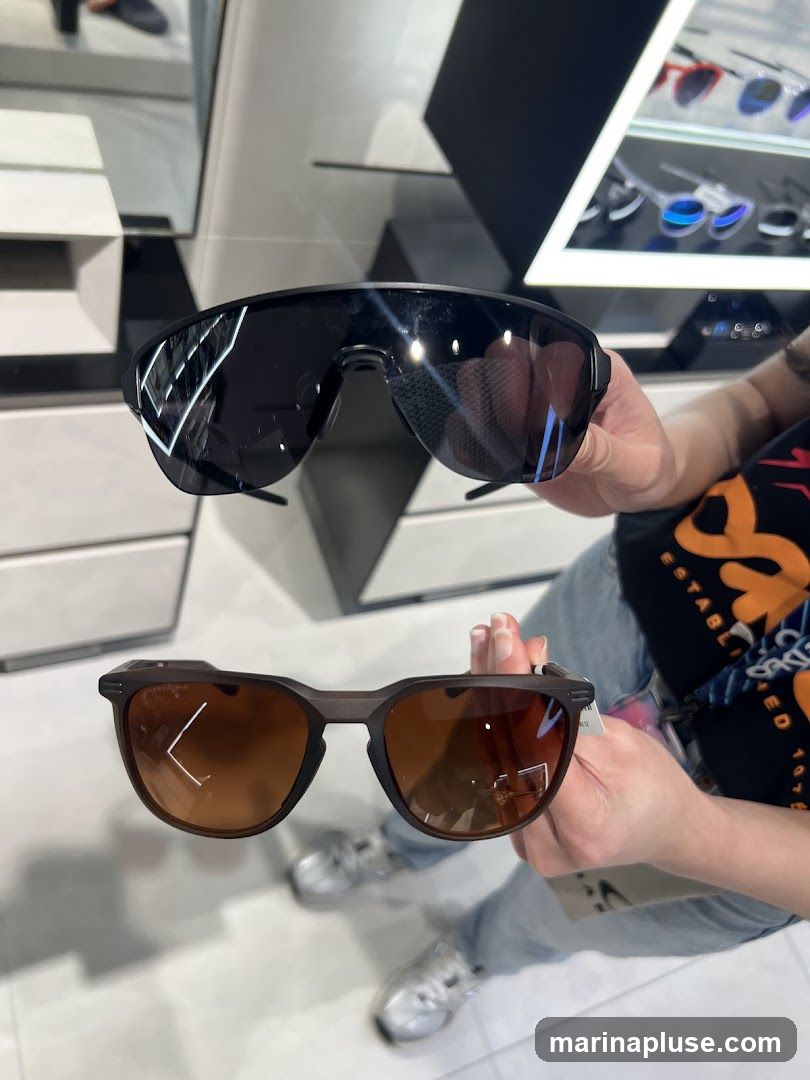 A person is holding up two pairs of sunglasses in a store. The top pair is black with dark lenses, and the bottom pair has a brown frame and brown lenses. In the background, there are displays of other sunglasses and store fixtures.