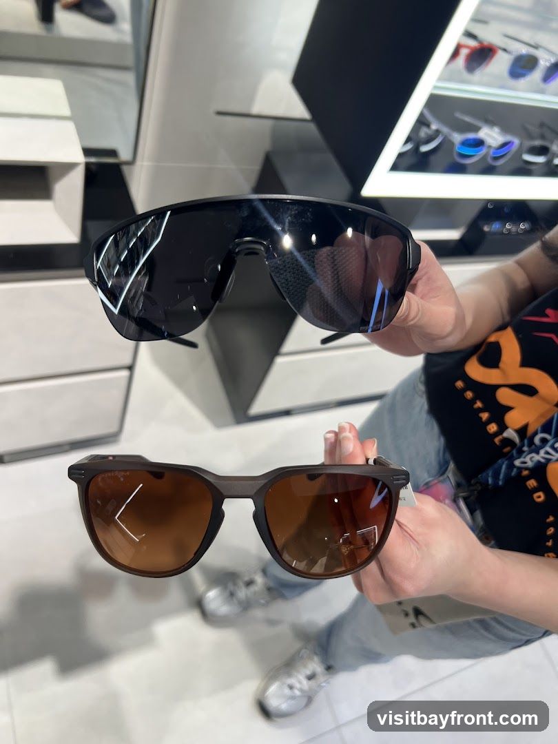 A person is holding up two pairs of sunglasses in a store. The top pair is black with dark lenses, and the bottom pair has a brown frame and brown lenses. In the background, there are displays of other sunglasses and store fixtures.
