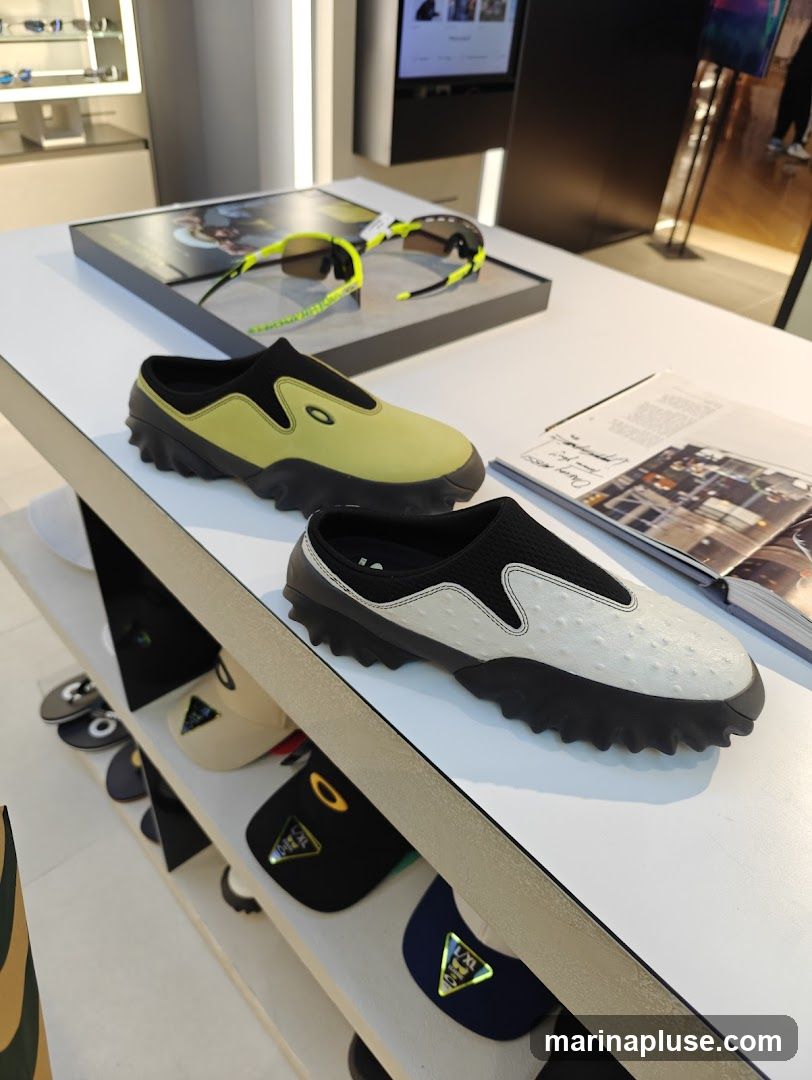 A display of Oakley footwear and sunglasses in a retail store. Two pairs of slip-on shoes are prominently featured on a white shelf. One shoe is lime green with black accents, and the other is white with a textured pattern and black accents. Both shoes have the Oakley logo. Behind the shoes, a pair of Oakley sunglasses with neon yellow accents is displayed on a black stand. Below the shoes, shelves display various Oakley caps and visors, some with the Oakley logo visible. In the background, a digital screen shows images, and a magazine or catalog lies open on the shelf.