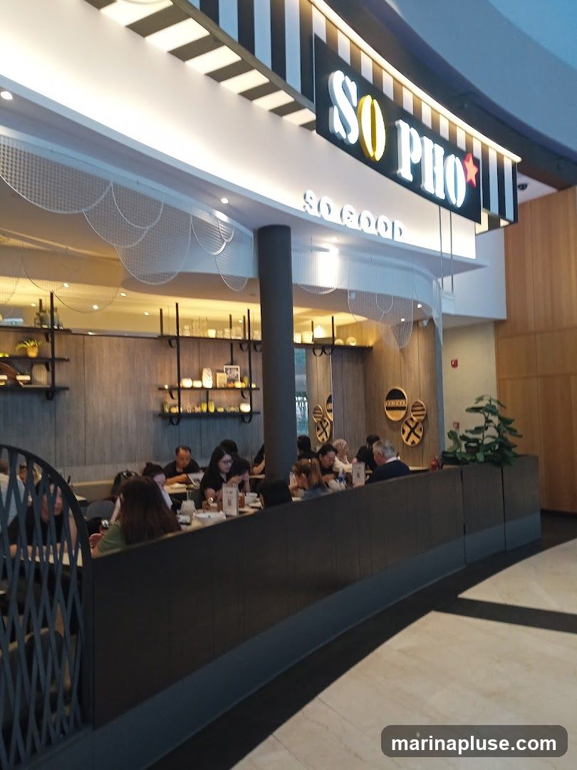 A view of the interior of a restaurant with diners seated at tables. The restaurant has a modern design with wooden accents and a prominent sign that reads "SO PHO SO GOOD".