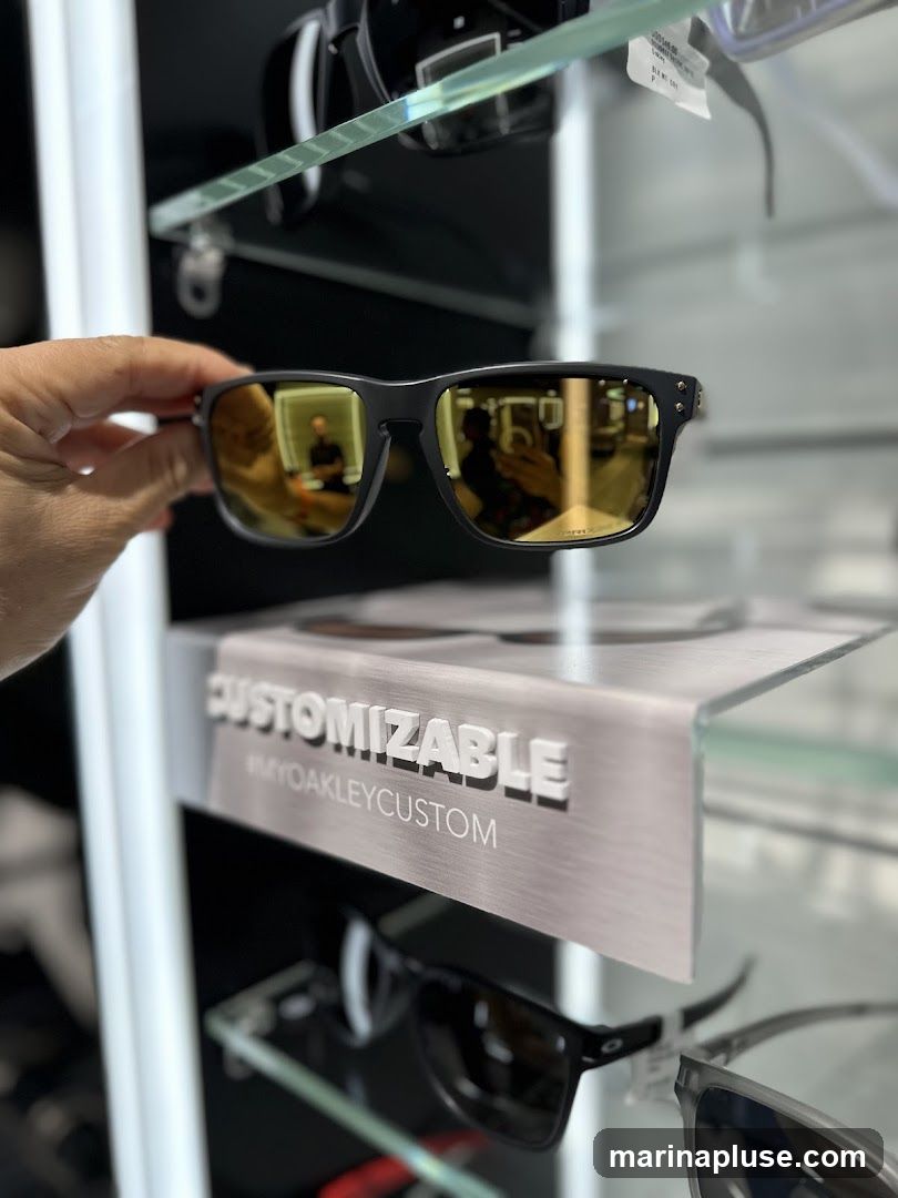 A hand holds a pair of black Oakley sunglasses with gold-tinted lenses. The sunglasses are displayed in a store setting, with other sunglasses visible on shelves behind. A sign reads "CUSTOMIZABLE #MYOAKLEYCUSTOM". The reflection in the sunglasses shows a person and the store interior.