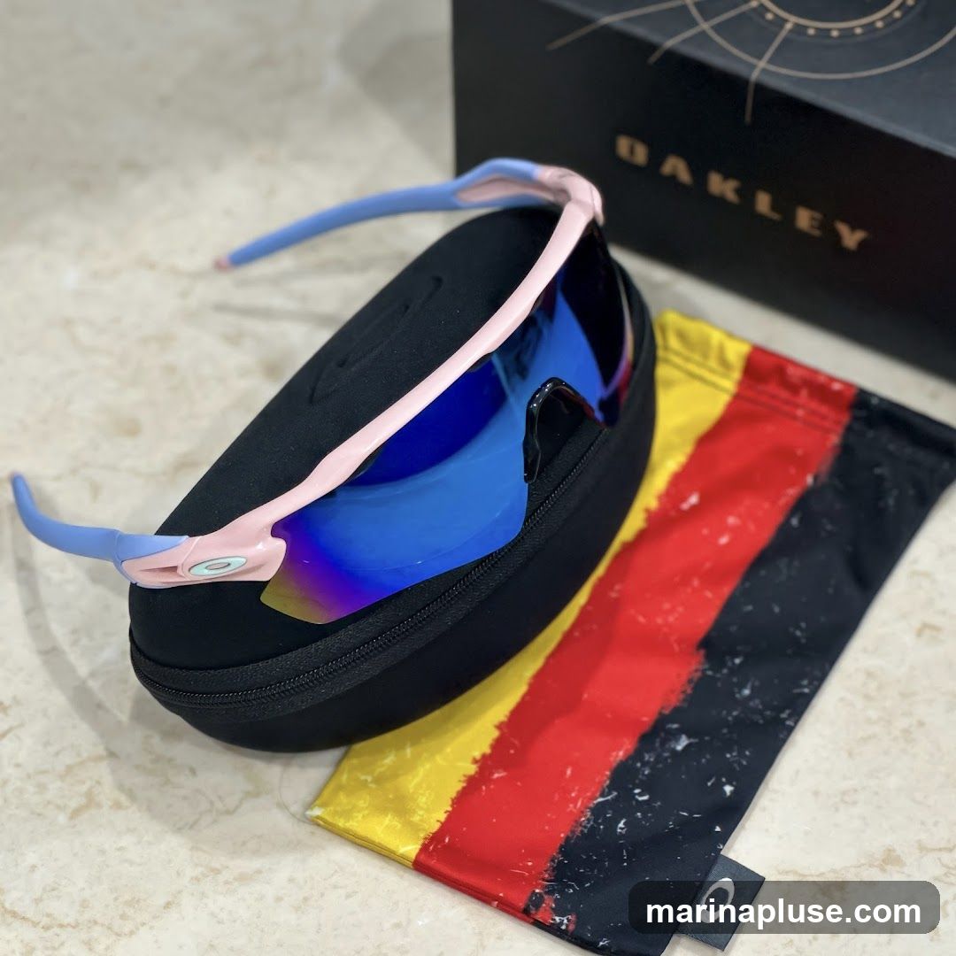 A pair of pink and light blue Oakley sunglasses with blue mirrored lenses rests on a black protective case. Next to the case is a soft pouch with a distressed German flag design. In the background, a black Oakley box is visible.