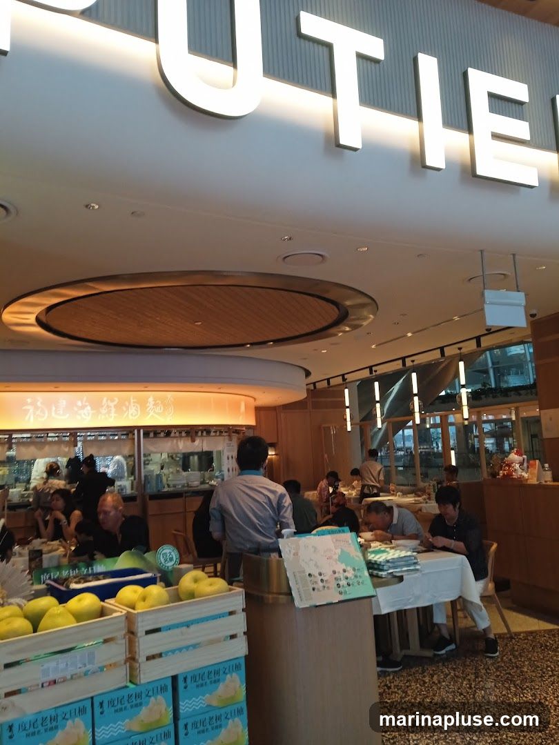 The image shows the interior of a restaurant with customers seated at tables. Large, illuminated letters spelling out "TIE" are visible on the ceiling. In the foreground, crates of fruit are displayed. A menu or informational pamphlet is on a table. The background shows the kitchen area and more dining tables.