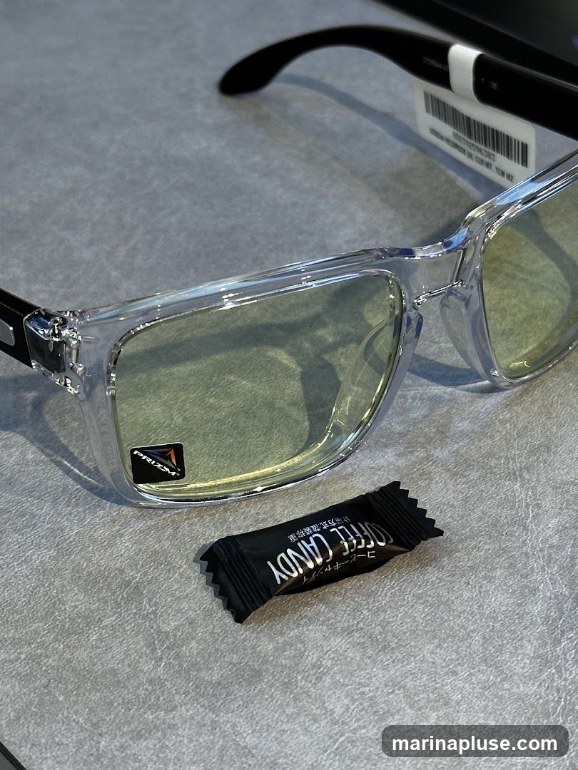 A pair of clear-framed Oakley sunglasses with yellow-tinted lenses rests on a gray surface. A small, black wrapper for "COFFEE CANDY" is placed in front of the sunglasses. A tag with product information is attached to the arm of the sunglasses.