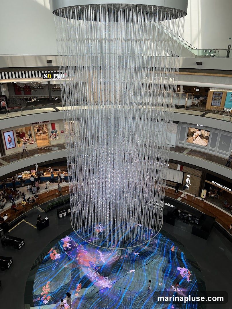 The image shows the interior of a modern shopping mall. A large, elaborate chandelier hangs from the ceiling, dominating the center of the frame. Below, the floor features a vibrant digital display with animated aquatic scenes and floral motifs. Several levels of the mall are visible, with various shops and escalators. Signs for businesses like 'SO PHO', 'MIKIHOUSE', 'PUTIEN', 'Dior', and 'STARBUCKS' are discernible on the upper levels. People are seen browsing shops and walking around the mall.