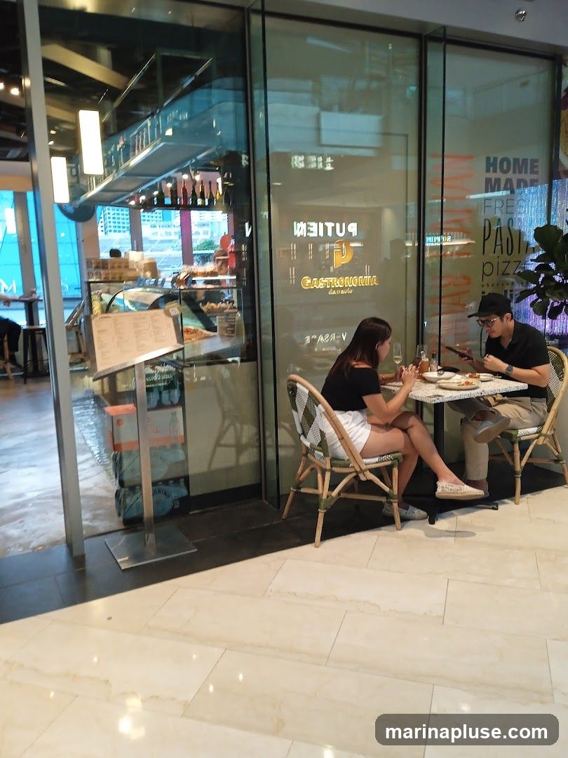 A couple dines at a table outside a restaurant with a glass facade. The restaurant's name, "GASTRONOMIA," is visible on the glass. Inside, a display case shows food items, and a menu stand is placed near the entrance.