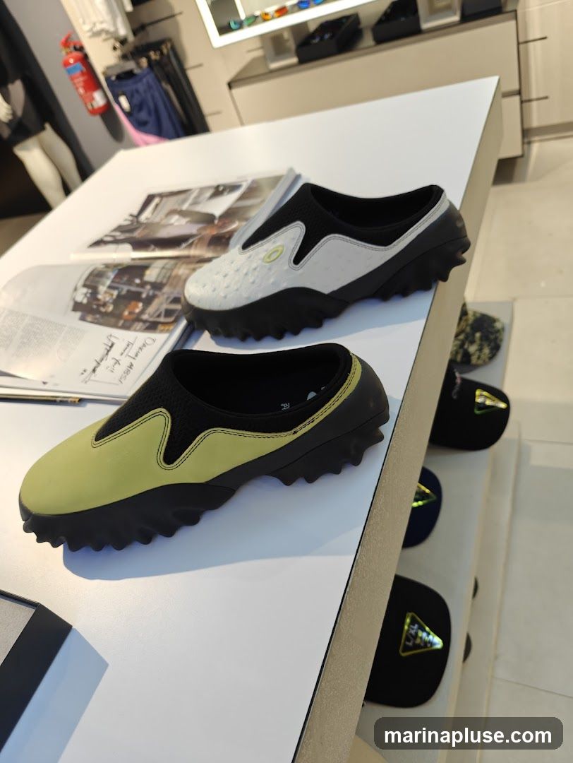 A display of Oakley shoes on a white table in a store. Two pairs of slip-on shoes are prominently featured in the foreground, one white with a textured pattern and black accents, and another in a light yellow with black accents. Behind them, on shelves, are several black caps with triangular logos. The background shows clothing racks with apparel and a display case with sunglasses.