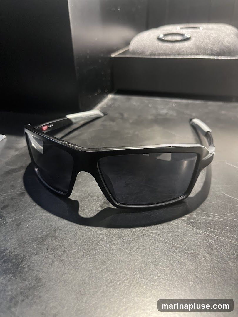 A pair of black Oakley sunglasses with dark lenses rests on a textured surface. A black case with the Oakley logo is visible in the background.