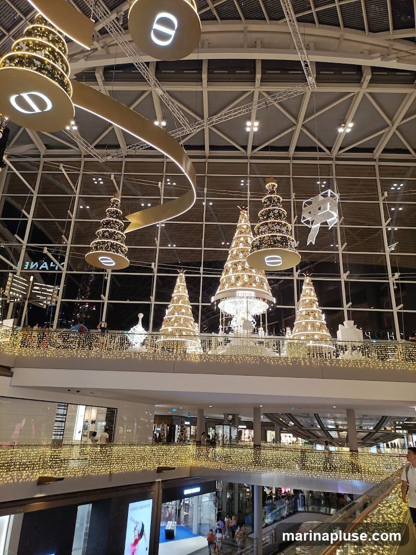 A festive indoor scene with multiple levels of a shopping mall decorated for Christmas. Large, illuminated Christmas trees are suspended from the ceiling, along with decorative ribbons and a large bow. The railings of each level are adorned with strings of warm, golden lights. Shops are visible on the lower levels, with some shoppers browsing. The overall ambiance is bright and festive.