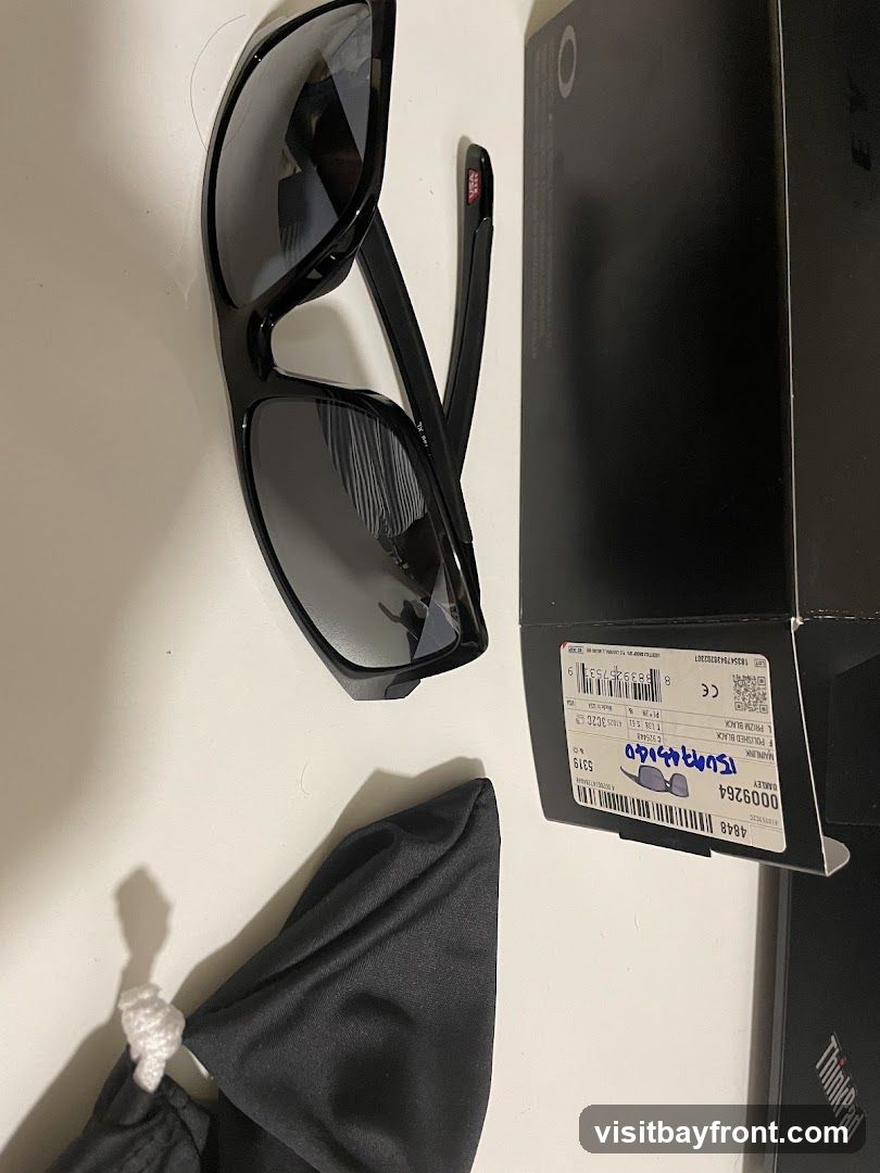 A pair of black Oakley sunglasses with polarized lenses is displayed next to its original packaging and a soft cleaning cloth. The box has a label with product details including the model name "MAINLINK F POLISHED BLACK" and a barcode.