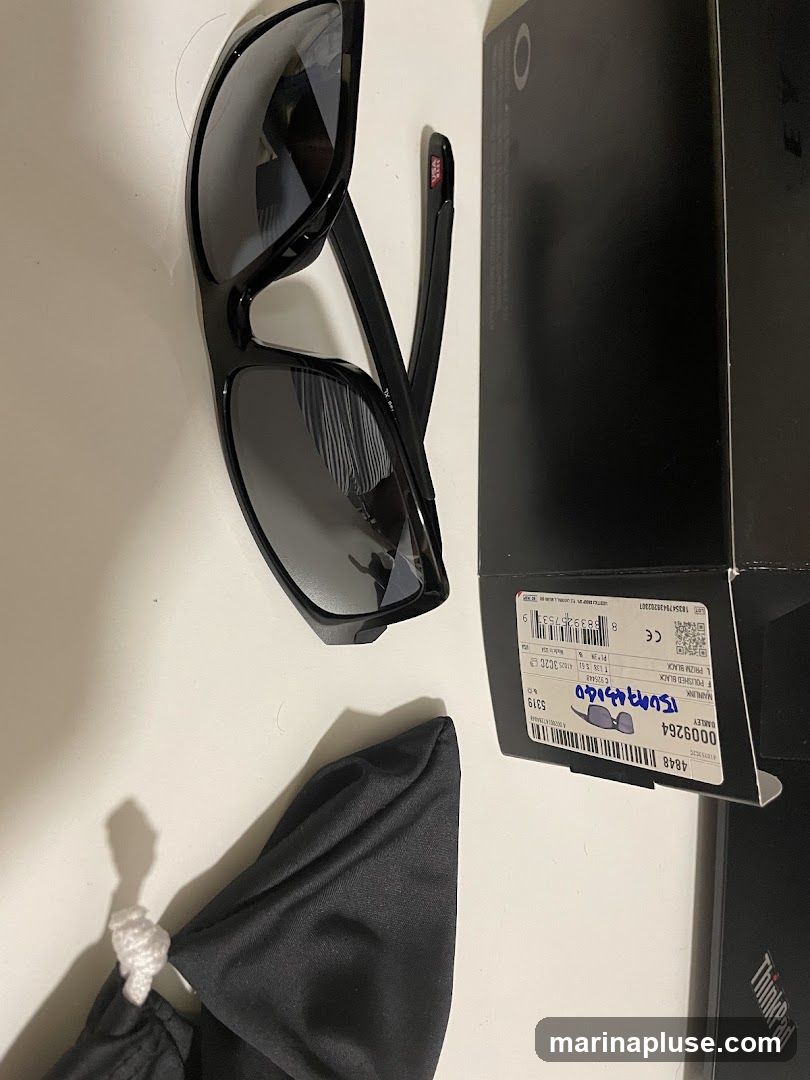 A pair of black Oakley sunglasses with polarized lenses is displayed next to its original packaging and a soft cleaning cloth. The box has a label with product details including the model name "MAINLINK F POLISHED BLACK" and a barcode.