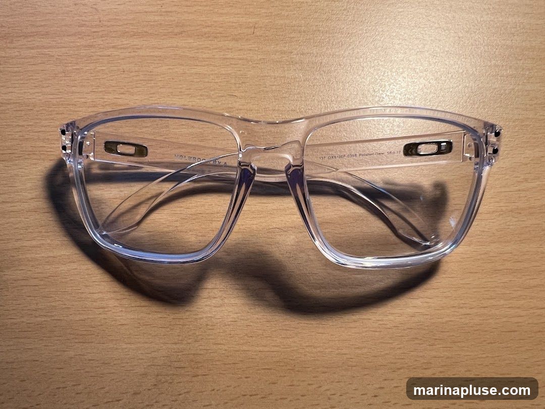 A pair of clear frame eyeglasses with the Oakley logo on the temples is shown on a wooden surface. The model number "137 OX8100F-0358 Polished Clear 58017" is visible on the inside of the right temple.